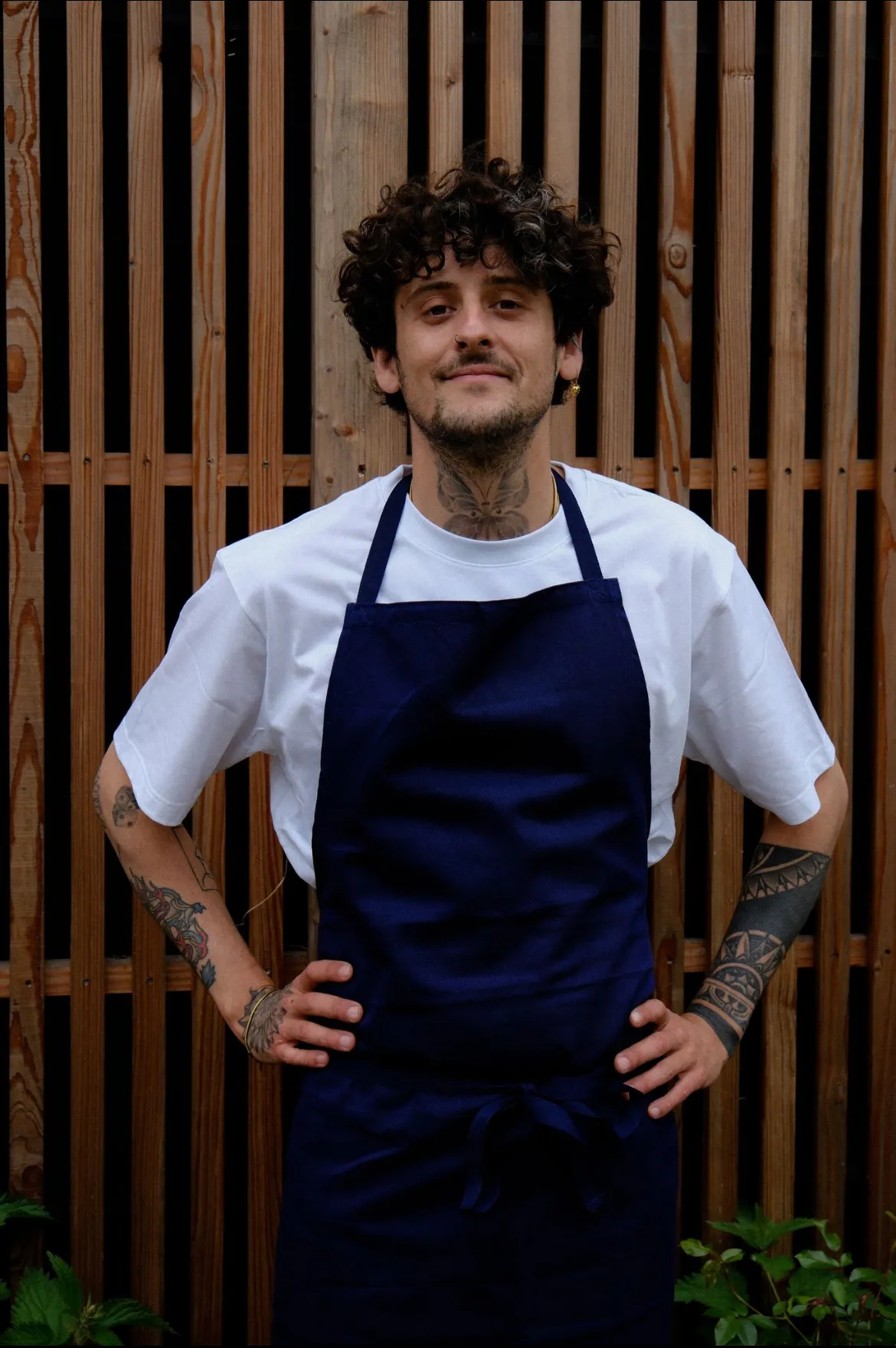 Alexandre L’Horphelin en tablier bleu, mains sur les hanches, devant une palissade en bois